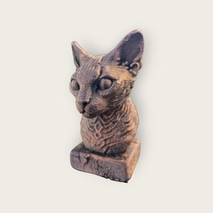 Puede incluir: Busto de gato en bronce. La escultura presenta una cabeza de gato detallada con ojos grandes y orejas prominentes. El busto está montado sobre una base rectangular, con una superficie texturizada. Diseño vintage.