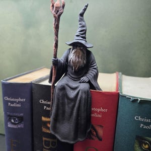 Könnte beinhalten: Eine 3D-gedruckte schwarze Zaubererfigur mit langem weißem Bart und spitzem Hut, die einen hohen braunen Stab mit Kristallspitze hält. Die Figur sitzt auf dem Rand eines roten Buches, platziert zwischen anderen Büchern von Christopher Paolini.