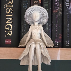May include: A detailed, off-white fairy figurine with a mushroom cap-like headpiece and flowing gown. The fairy is seated, with outstretched arms and delicate wings. The figurine is placed on a wooden shelf in front of a row of books.