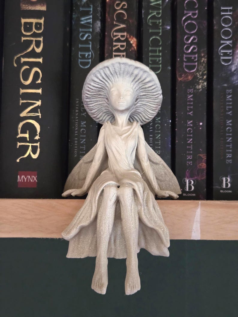 May include: A detailed, off-white fairy figurine with a mushroom cap-like headpiece and flowing gown. The fairy is seated, with outstretched arms and delicate wings. The figurine is placed on a wooden shelf in front of a row of books.
