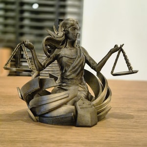 Könnte beinhalten: Eine bronzene Statue einer Frau, die in jeder Hand eine Waage hält. Die Frau sitzt auf einem Sockel aus abstrakten, geometrischen Formen. Die Statue ist eine Darstellung der Gerechtigkeit.