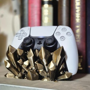 Op de afbeelding: Een witte PlayStation 5-controller staat op een gouden, kristalvormige standaard. De standaard is gemaakt van meerdere gouden kristallen die in een cluster zijn gerangschikt.