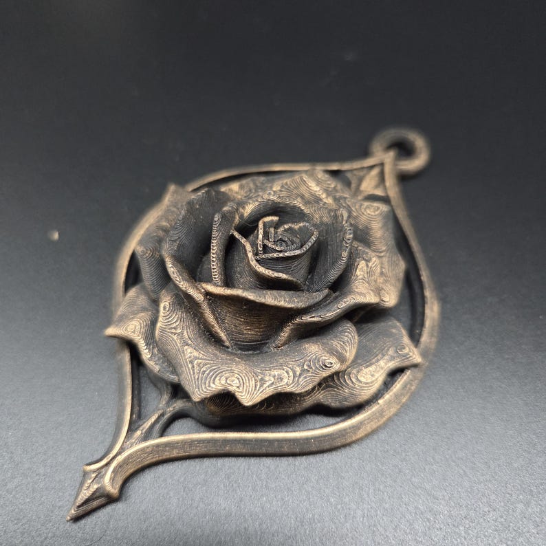 May include: A bronze-colored pendant featuring a detailed, layered rose bloom centered within a pointed, leaf-shaped frame. The metal surface displays a distinct, fine-lined texture throughout the petals and the surrounding decorative border.