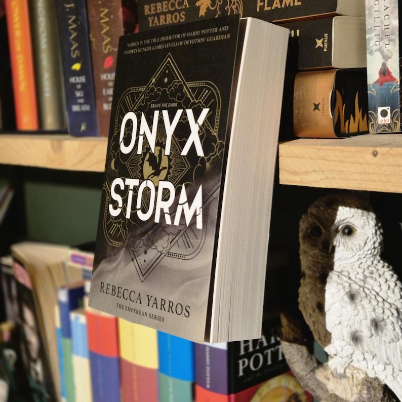 May include: A black and gold book titled "Onyx Storm" by Rebecca Yarros. The book is part of the Empyrean series and features a dark, fantasy-themed cover with a stylized design.