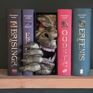 Op de afbeelding: Een unieke boekensteun met een monstergezicht en klauw, geplaatst tussen boeken. De boeken hebben titels als "Brsingr" en "Eragon" van Christopher Paolini. De boekensteun is een gedetailleerde, getextureerde sculptuur.