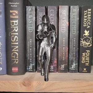 May include: A silver-colored figurine of a knight reading a book, positioned in front of a row of books. The books have titles like "Twisted" and "Fourth Wing." The figurine is detailed, with visible armor and a helmet. The books are on a wooden shelf.