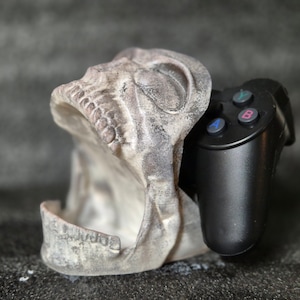 Può includere: Un supporto bianco a forma di teschio con un controller per videogiochi nero all'interno. Il controller ha pulsanti etichettati A, B e Y.