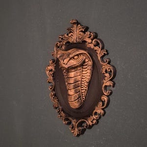 Könnte beinhalten: Bronzefarbene Cobra-Skulptur in einem verzierten, ovalen Rahmen. Die Cobra ist zentriert, mit detaillierten Schuppen und einem fokussierten Blick. Der Rahmen hat aufwendige Schnörkel und einen dunkelbraunen Hintergrund.