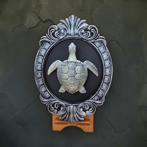Op de afbeelding: Een decoratieve wanddecoratie met een zilverkleurige, sierlijke ovale lijst. Binnenin toont een zwarte achtergrond een gedetailleerde, driedimensionale schildpadsculptuur. Het stuk is gemonteerd op een houten voet.