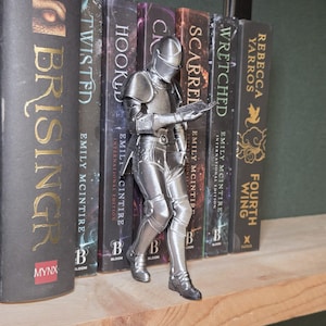 May include: A silver knight figurine reading a book, positioned among a row of hardcover books. The knight is detailed with armor and a helmet. The books have titles like "Twisted" and "Fourth Wing". The scene is set on a wooden shelf.