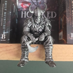 Op de afbeelding: Een zilverkleurig beeldje van een zittende Vikingkrijger met een gehoornde helm en schouderharnas met bonttextuur. Het figuur heeft een lange baard en houdt een strijdbijl vast, met gedetailleerde gelaagde kleding en laarzen zichtbaar op het personage.