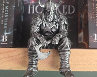 Könnte beinhalten: Eine silberfarbene Figur eines sitzenden Wikingerkriegers mit gehörntem Helm und fellartiger Schulterpanzerung. Die Figur hat einen langen Bart und hält eine Streitaxt in der Hand, wobei detaillierte geschichtete Kleidung und Stiefel sichtbar sind.