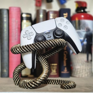 Op de afbeelding: Een witte PlayStation 5-controller wordt getoond op een gouden, touwachtige standaard. De controller rust op de standaard met de voorkant naar de kijker gericht. De standaard staat op een houten oppervlak.