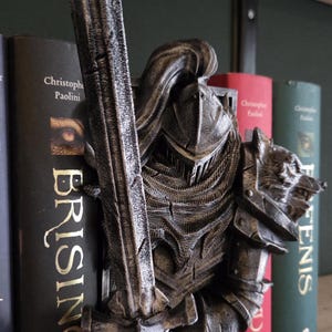 May include: A detailed, metallic-looking knight figurine with a sword, positioned between books on a shelf. The knight is in full armor, with a helmet and a wolf's head on the shoulder. The books have gold lettering.