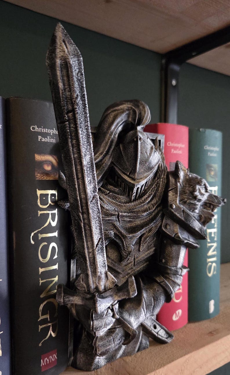 May include: A detailed, metallic-looking knight figurine with a sword, positioned between books on a shelf. The knight is in full armor, with a helmet and a wolf's head on the shoulder. The books have gold lettering.