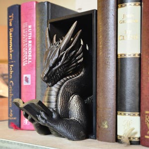 May include: A black dragon bookend holding a book. The dragon is reading the book with its left hand. The bookend is made of a textured material and has a detailed design. The dragon is positioned between two bookshelves with books on each side.