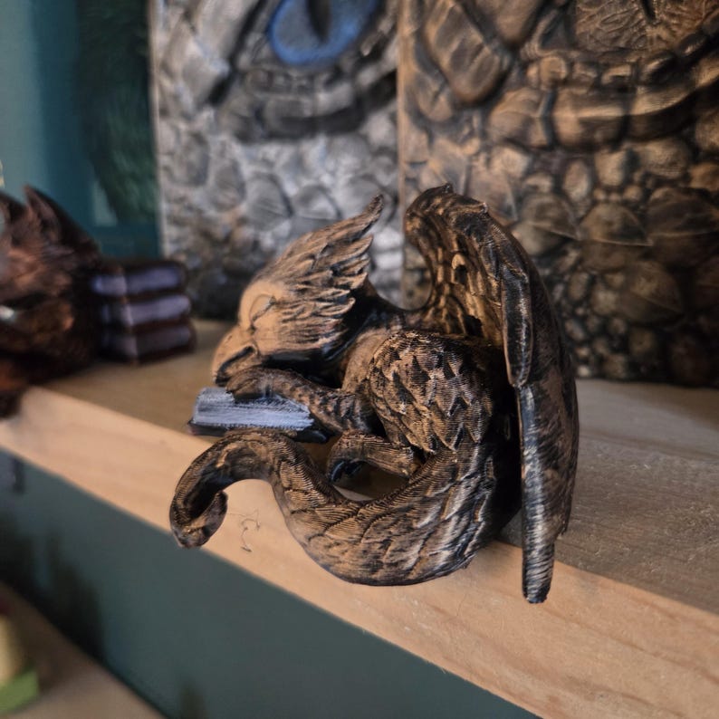 May include: A detailed sculpture of a sleeping griffin, featuring a bird-like head, feathered wings, and a serpentine tail. The griffin is resting on a small, rectangular base. The sculpture is primarily brown with darker shading, giving it a textured appearance.