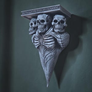 Peut inclure: Une étagère murale grise de style gothique avec trois crânes soutenant une étagère rectangulaire. L'étagère présente un design détaillé et texturé, et les crânes sont sculptés avec des détails squelettiques.