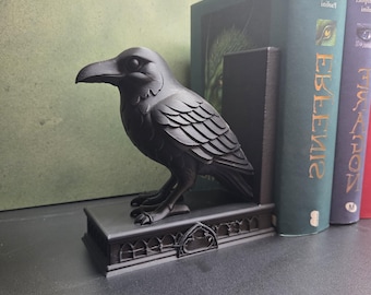 May include: A matte black bookend featuring a sculpted raven perched on a rectangular base. The base is decorated with a series of arched, gothic-style window cutouts along the front edge. The raven is depicted in profile, facing left.