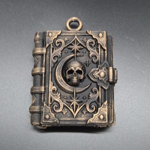 May include: A rectangular pendant shaped like a book with a black and bronze finish. The cover features a central skull, a crescent moon, and star motifs surrounded by ornate scrollwork. A small circular loop is attached to the top edge.