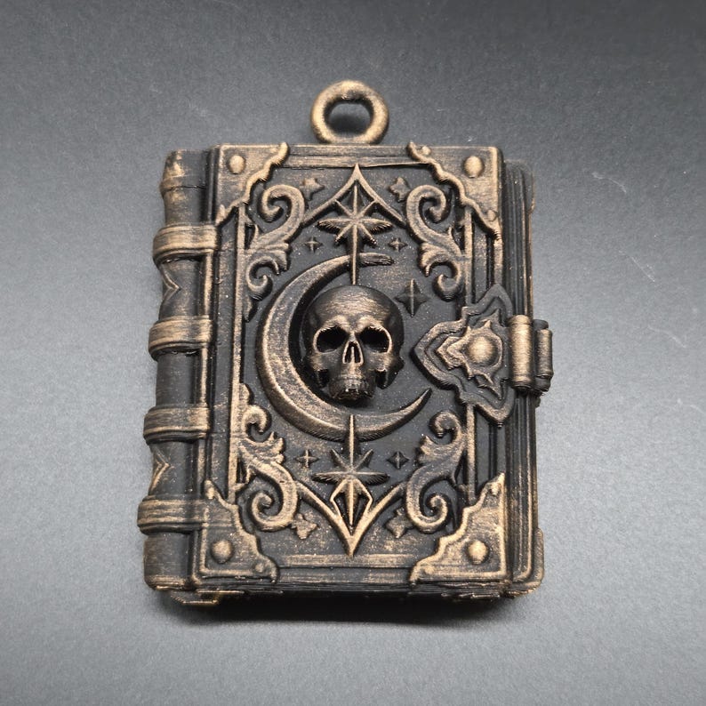 May include: A rectangular pendant shaped like a book with a black and bronze finish. The cover features a central skull, a crescent moon, and star motifs surrounded by ornate scrollwork. A small circular loop is attached to the top edge.