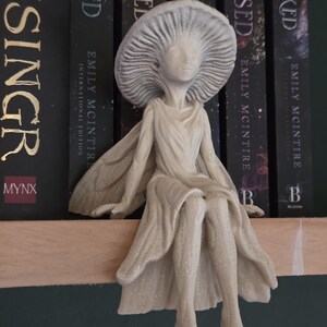 May include: A beige figurine of a seated fairy with a mushroom-shaped hat. The fairy is wearing a flowing gown and has outstretched arms. The figurine is placed in front of a row of books with black spines.