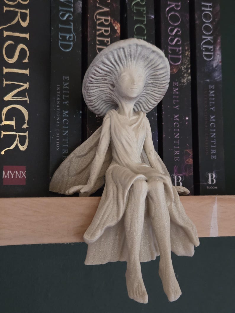 May include: A beige figurine of a seated fairy with a mushroom-shaped hat. The fairy is wearing a flowing gown and has outstretched arms. The figurine is placed in front of a row of books with black spines.