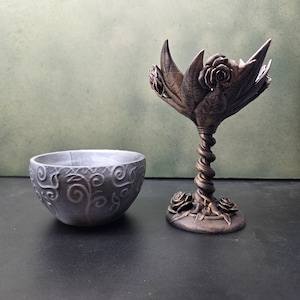 Könnte beinhalten: Eine silberfarbene Schale mit geprägten Wirbelmustern neben einem bronzefarbenen Kelch mit gedrehtem Stiel, Rosenmotiven am Fuß und einer Schale in Form einer blühenden Blume mit spitzen Blütenblättern und zentralen Rosendetails.