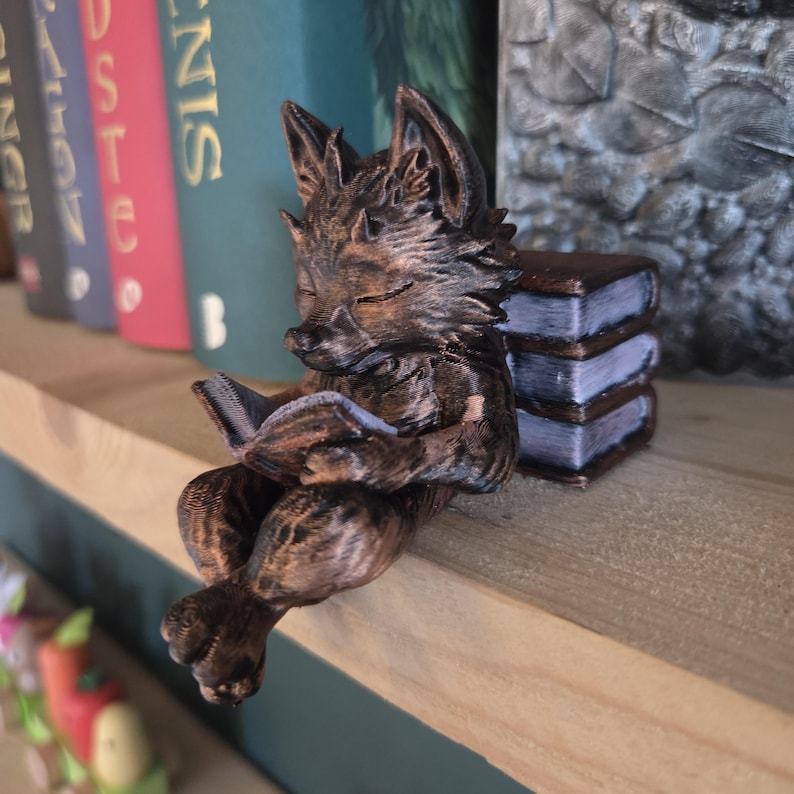 May include: A brown fox figurine reading a book, perched on a wooden shelf. The fox is detailed with textured fur and closed eyes, holding a blue-paged book. Beside it is a stack of three miniature books. The background includes colorful book spines.