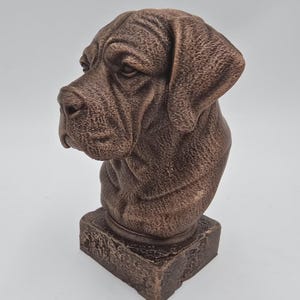 May include: A brown dog bust sculpture. The sculpture shows the head and upper chest of a dog with detailed facial features, including wrinkles and folds. The bust is mounted on a rectangular base.