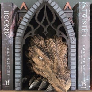 May include: A bookend featuring a sculpted dragon head with textured tan and brown scales emerging from a dark gray gothic-style arched stone structure. The dragon has visible claws resting at the base of the archway, positioned between two books.