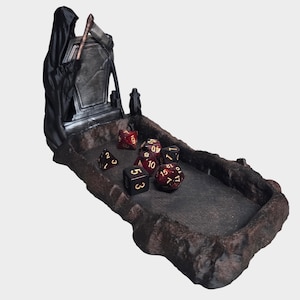 May include: A themed dice rolling tray featuring a grim reaper figure and tombstone design. The tray is dark grey with a textured surface, and several dice are scattered inside. The reaper holds a scythe, adding to the gothic aesthetic.