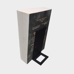 May include: A hardcover book with a black cover and white pages is held upright by a black metal bookend. The bookend has a rectangular base and a vertical support. Text is visible on the book cover.