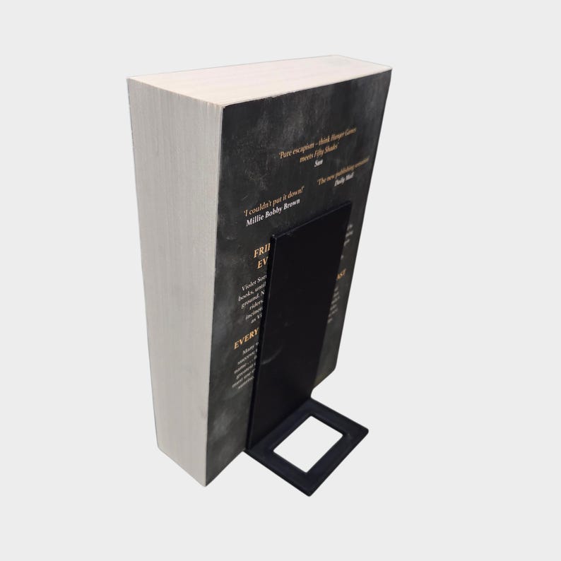 May include: A hardcover book with a black cover and white pages is held upright by a black metal bookend. The bookend has a rectangular base and a vertical support. Text is visible on the book cover.