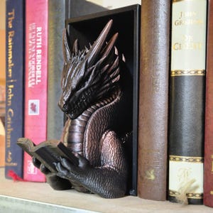 May include: A black dragon bookend with a detailed, textured design. The dragon is holding a book in its claws and is positioned as if it is reading. The bookend is made of a dark, metallic material and is a great addition to any bookshelf.
