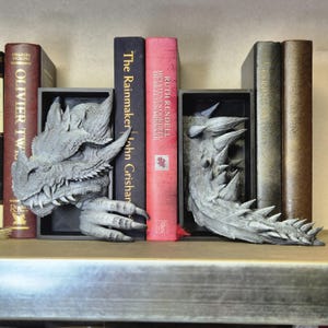 Könnte beinhalten: Ein Paar graue Drachen-Buchstützen, die Bücher in einem Regal halten. Die Buchstützen sind aus einem strukturierten Material gefertigt und haben ein realistisches Drachen-Design. Die Bücher im Regal sind "The Rainmaker" von John Grisham und "The Stone Order" von Ruth Rendell.