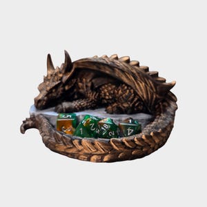 Könnte beinhalten: Eine bronzefarbene Drachenfigur, die sich um einen grauen Steinsockel windet und detaillierte Schuppen sowie Hörner aufweist. Der Schwanz des Drachen umschließt eine kleine Sammlung mehrseitiger Würfel in Grün- und Orangetönen mit weißen Zahlen.