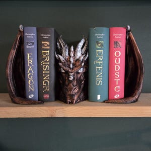 Könnte beinhalten: Bronzefarbene Drachenkopf-Buchstützen mit ausgebreiteten Flügeln. Der Drachenkopf befindet sich zwischen den Flügeln, mit vier Büchern dazwischen. Die Bücher tragen die Titel "Eragon", "Brisingr", "Erfenis" und "Oudste".