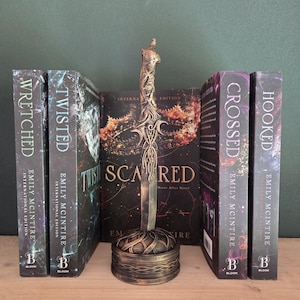 Op de afbeelding: Een decoratief bronskleurig dolkbeeldhouwwerk met een versierd handvat en een ronde voet, geplaatst voor vijf boeken getiteld Wretched, Twisted, Scared, Crossed en Hooked van Emily McIntire, met donkere, hemelse omslagillustraties.