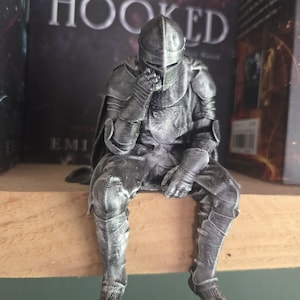 May include: A silver-toned 3D printed figurine of a knight in full plate armour, depicted in a seated, pensive pose with one hand resting near the helmet. The figure features visible layer lines from the printing process and a weathered finish.