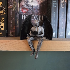 Peut inclure: Une figurine argentée représentant une créature ailée assise sur un rebord en train de lire un livre. La figurine possède de grandes ailes sombres et texturées déployées, vêtue d'une chemise à col et d'un pantalon, les jambes pendantes.