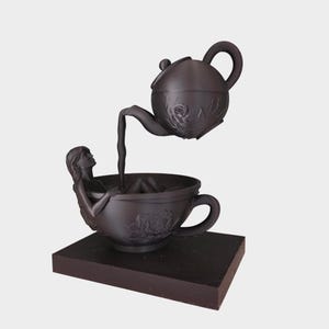 Op de afbeelding: Een donkerbruine sculptuur van een vrouw in een theekopje, met een theepot die vloeistof giet. De theekop en theepot zijn versierd met bloemmotieven, en de sculptuur staat op een rechthoekige basis.