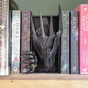 Op de afbeelding: Een zwarte boekensteun in gotische stijl met een gestileerde drakenklauw met een gouden ring en een gedetailleerde drakenkop. De boekensteun staat tussen een rij boeken op een houten plank.