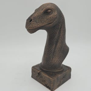 Pode incluir: Escultura de cabeça de dinossauro em tom bronze. A escultura apresenta uma representação detalhada da cabeça e pescoço de um dinossauro, montada numa base quadrada. O dinossauro tem uma superfície texturizada e é um objeto decorativo.