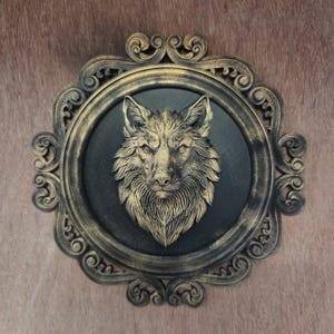 Könnte beinhalten: Eine kreisförmige Wandplakette mit einem dreidimensionalen, goldfarbenen Wolfskopf-Relief auf schwarzem Hintergrund. Das Stück ist in einen verzierten, goldfarbenen Rahmen mit Schnörkelmuster und Antik-Finish eingefasst, ein dekoratives Wandobjekt.