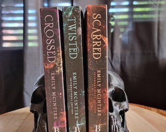 Puede incluir: Tres libros titulados "Crossed", "Twisted" y "Scarred" de Emily McIntire, sostenidos por un sujetalibros de calavera negra. Las lomos de los libros son oscuros con acentos coloridos. Los libros son parte de la serie "International Edition".