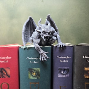 Pode incluir: Uma estatueta de gárgula cinzenta com asas, chifres e dentes afiados pousada sobre uma fila de livros. Os livros são da autoria de Christopher Paolini e apresentam ilustrações de olhos de dragão nas lombadas em vermelho, verde, roxo e preto.