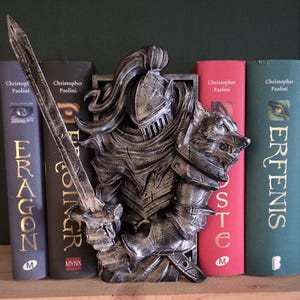 Op de afbeelding: Een zilverkleurige ridder boekensteun met een wolfskop aan de rechterkant, die een zwaard vasthoudt. De boekensteun staat tussen verschillende boeken met titels van Christopher Paolini. De boeken zijn in verschillende kleuren, waaronder donkerblauw, rood en groen.