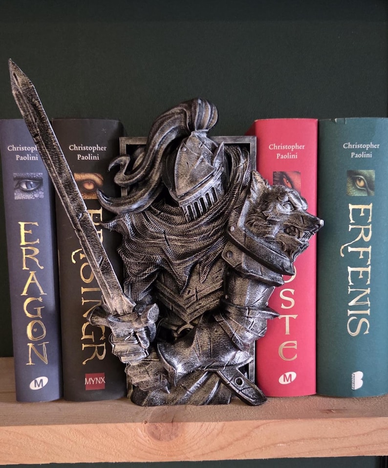 May include: A silver-colored knight bookend with a wolf head on the right side, holding a sword. The bookend is placed between several books with titles by Christopher Paolini. The books are in various colors, including dark blue, red, and green.