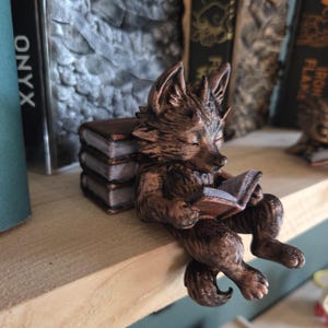 May include: A bronze-colored fox figurine reading a book, perched on a wooden shelf. The fox is sitting with its legs dangling, and a stack of miniature books is behind it. The word "ONYX" is visible on a book in the background.